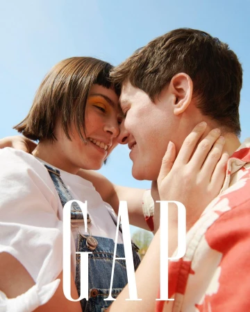 Smiling couple embracing outdoors in a close-up shot, wearing a white t-shirt, denim overalls, and a red patterned shirt, with the GAP logo.