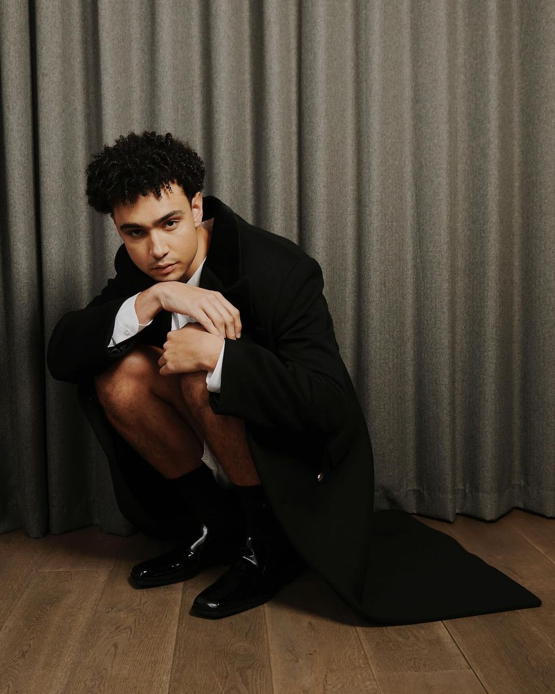 Young adult male with curly hair poses crouching in a black coat and shiny shoes in a studio setting with a grey curtain backdrop.