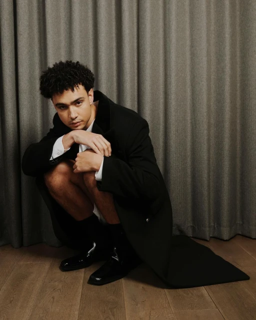 Young adult male with curly hair poses crouching in a black coat and shiny shoes in a studio setting with a grey curtain backdrop.