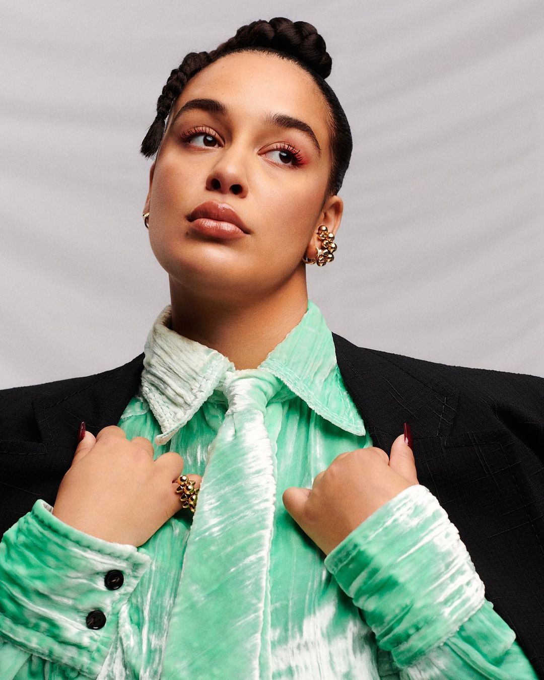 Adult woman in a black blazer and green crushed velvet shirt poses in a studio with soft lighting, wearing statement jewelry.