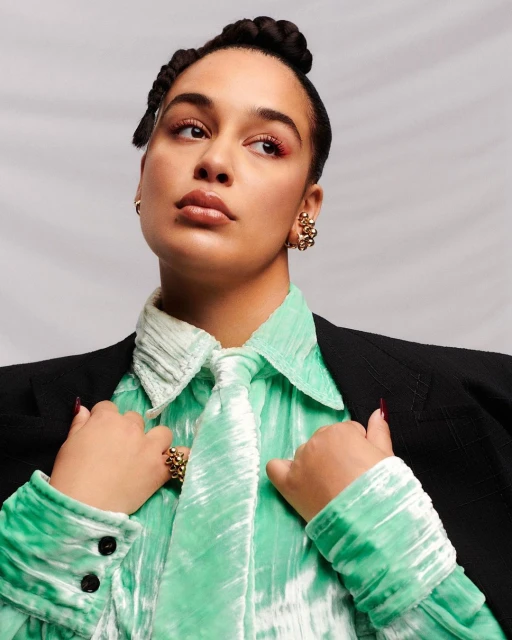 Adult woman in a black blazer and green crushed velvet shirt poses in a studio with soft lighting, wearing statement jewelry.