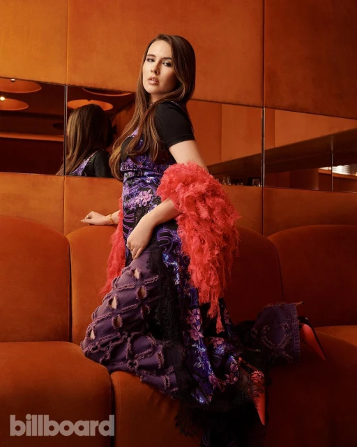 A woman in a floral dress and coral boa sits on an orange couch in an indoor setting, with the Billboard logo visible.