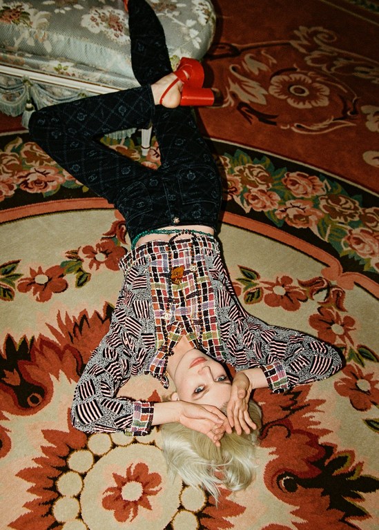 A person in patterned shirt and pants lies on an orange floral rug next to a decorative chair and is posing for a fashion editorial shot.