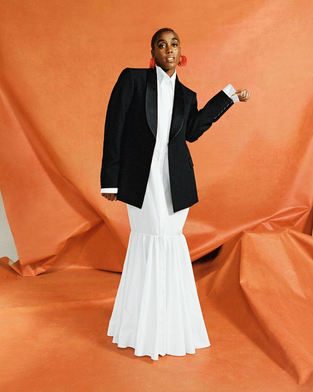 Woman in black blazer and white dress poses against an orange backdrop, wearing large red earrings and looking at the camera.