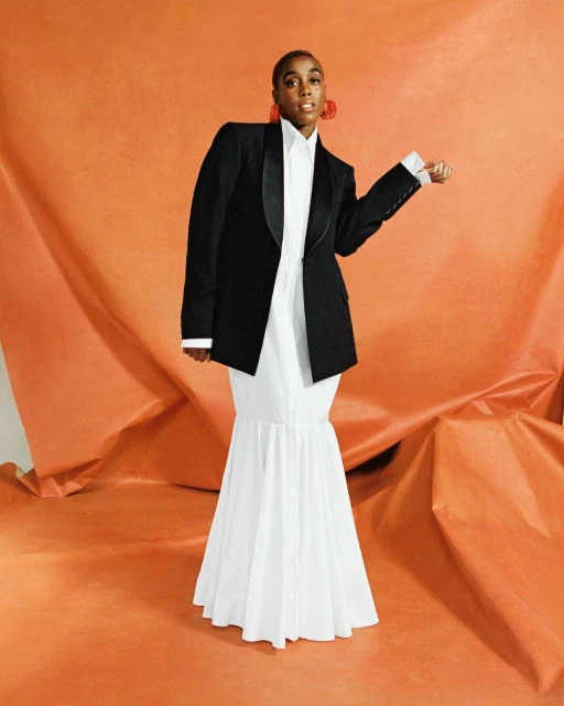 Woman in black blazer and white dress poses against an orange backdrop, wearing large red earrings and looking at the camera.