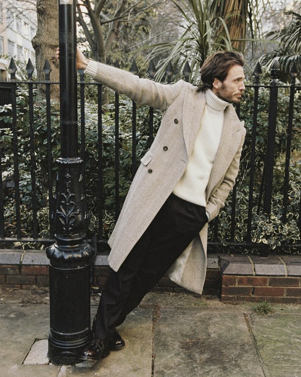 A stylish adult male with beard and coat poses next to a black lamp post in a leafy urban setting, wearing a beige coat and black trousers.