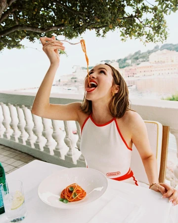 Woman in white halter top eating spaghetti from a fork outdoors with coastal town in background