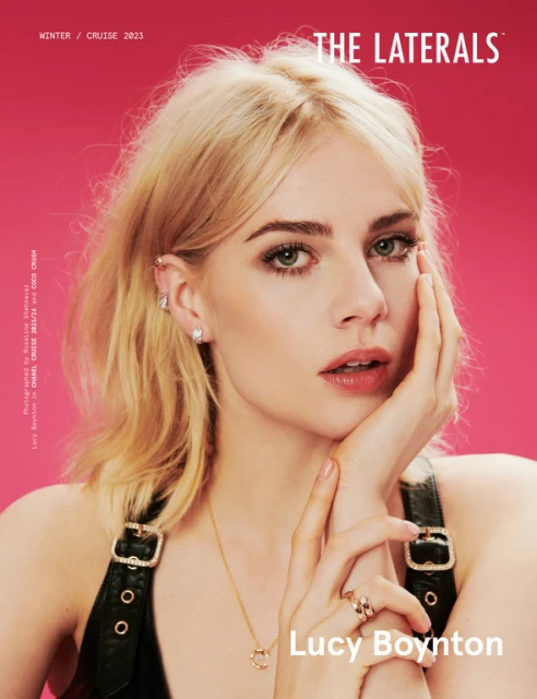 Close-up portrait of Lucy Boynton, with shoulder-length blonde hair, jewelry, and dark sleeveless top against a pink background.