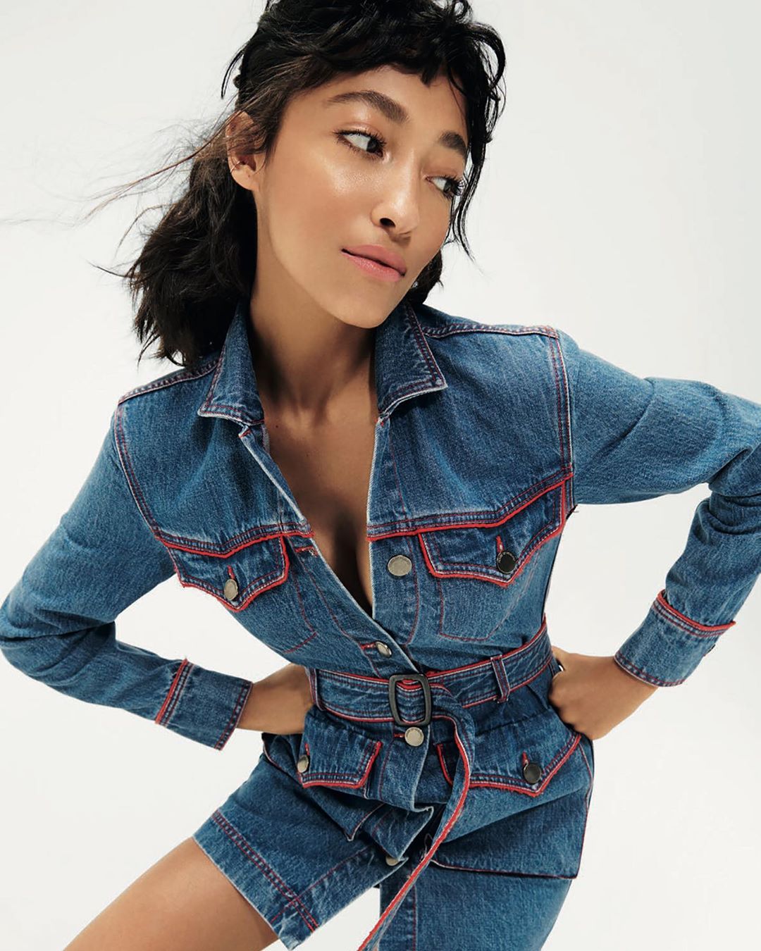 Portrait of an adult woman wearing a blue denim jacket dress with red trim on a seamless white backdrop in a studio shot.
