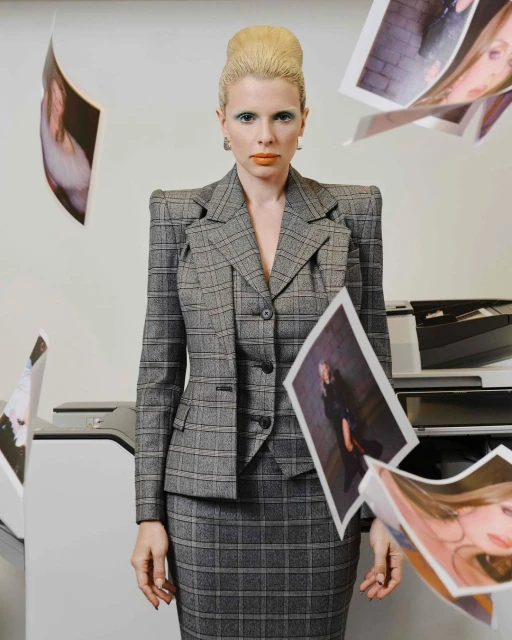 Adult female in a grey plaid suit stands in front of a printer with printed photos floating around her in a photo studio.