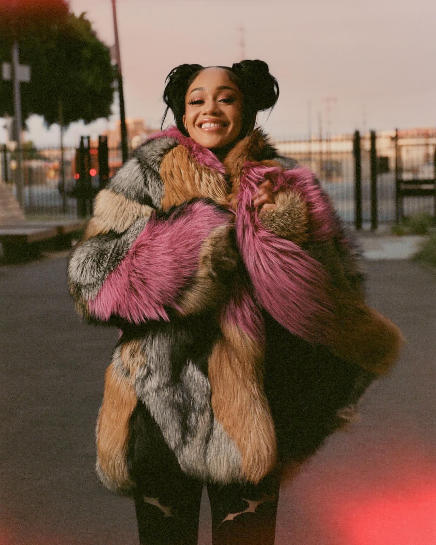 Smiling woman wearing a colorful fur coat in an urban setting with buildings and an orange sky; she has her hair in space buns.