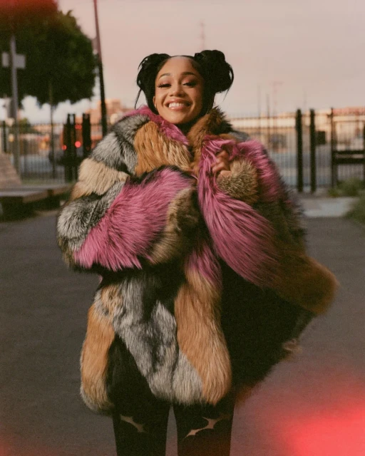Smiling woman wearing a colorful fur coat in an urban setting with buildings and an orange sky; she has her hair in space buns.