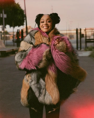 Smiling woman wearing a colorful fur coat in an urban setting with buildings and an orange sky; she has her hair in space buns.