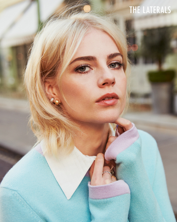Close-up portrait of a blonde woman with fair skin wearing a blue sweater with a white collar, gazing into the camera.