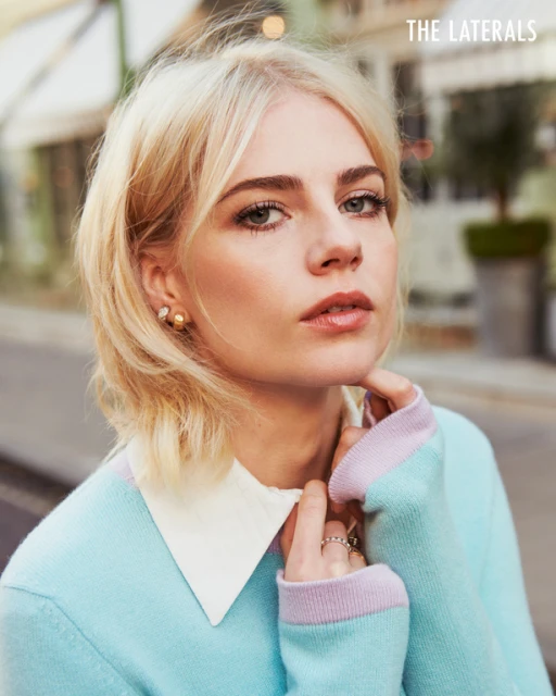 Close-up portrait of a blonde woman with fair skin wearing a blue sweater with a white collar, gazing into the camera.