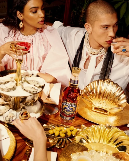 Two people at an elaborate table with oysters, liquor, and gold serving dishes, creating a scene of luxury and indulgence.