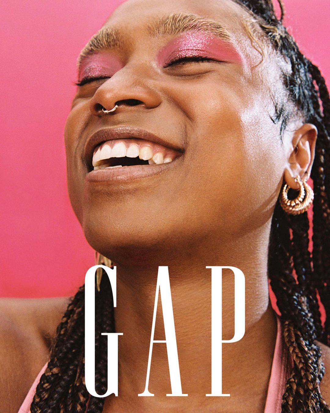 Close-up of a smiling adult woman with braided hair and visible makeup, wearing a hoop earring against a bright pink backdrop.