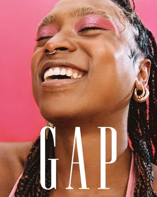 Close-up of a smiling adult woman with braided hair and visible makeup, wearing a hoop earring against a bright pink backdrop.