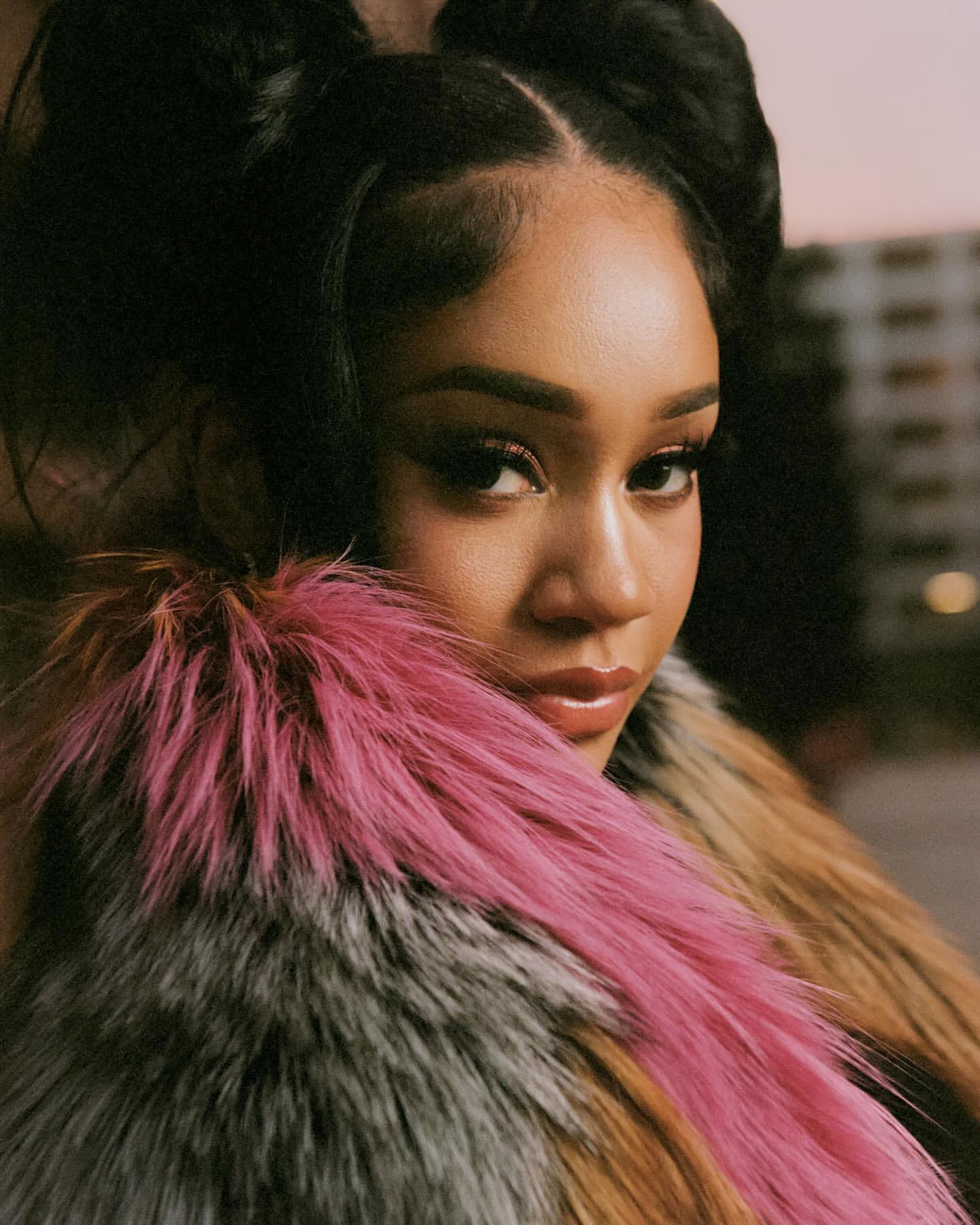 Close-up portrait of a woman with hair styled in space buns, wearing a multicolored fur shawl, against a blurred urban backdrop.