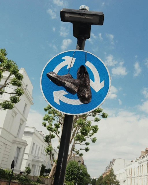 Pair of black dress shoes hanging from a roundabout sign, suspended by the laces on a sunny urban street with white buildings.