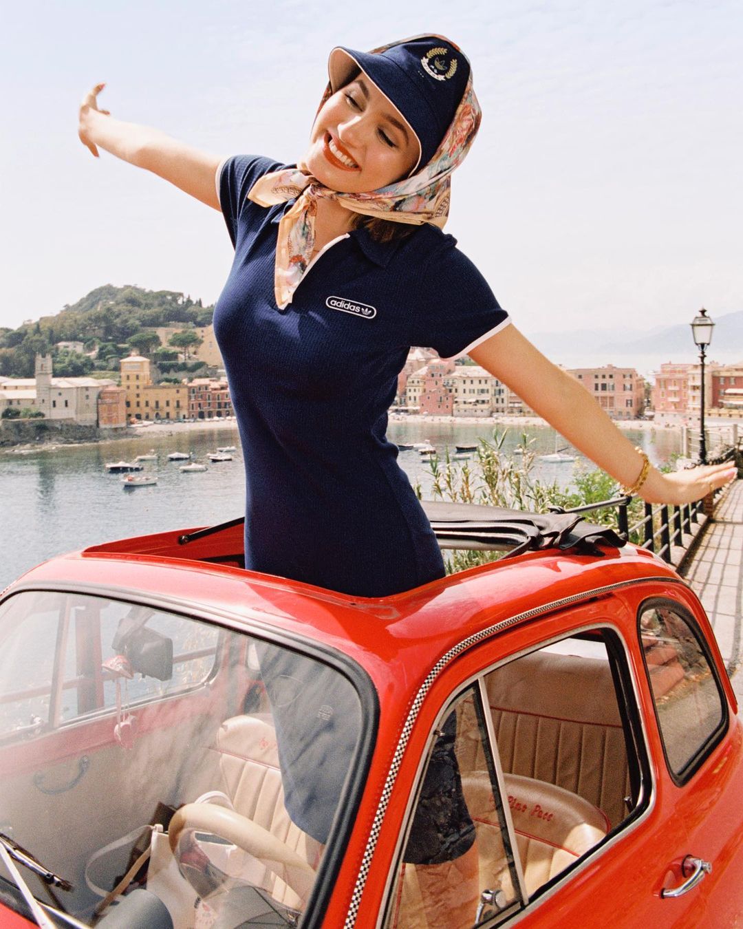 A happy woman in a vintage red car smiles with open arms, enjoying the Italian coastal view, wearing a cap and Adidas polo dress.