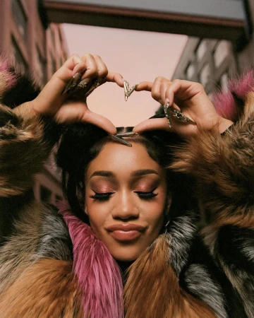 Close-up portrait of a woman making a heart sign with her hands, wearing a fluffy fur coat against an urban street background.