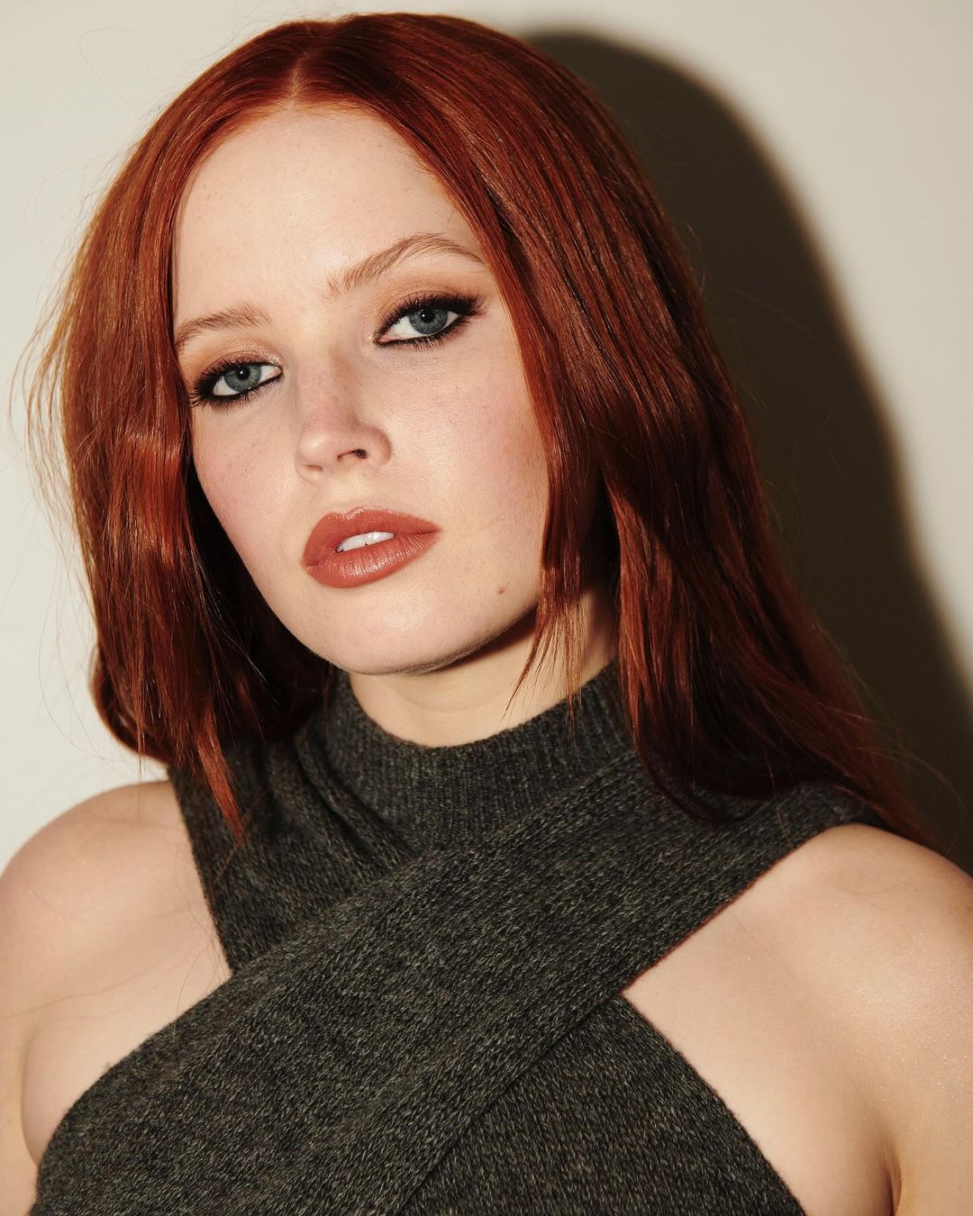 Portrait of a red-haired woman wearing a grey knit top against a light background, emphasizing her eye makeup and neutral lip color.