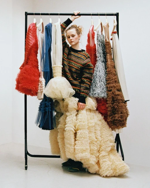 Adult female model stands in front of a clothing rack filled with dresses, wearing a patterned sweater and large tulle skirt in a studio.