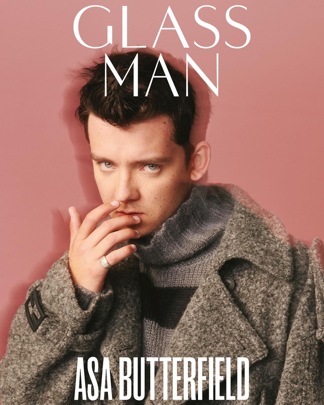 Portrait of Asa Butterfield in a grey coat, with 'Glass Man' magazine text, set against a pale pink background. Fashion and celebrity.