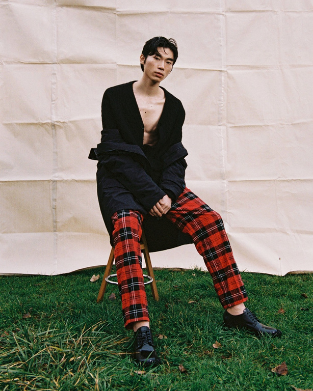 An Asian adult male fashion model sits on a stool in a photo studio wearing a black jacket, red plaid pants, and shiny black shoes.