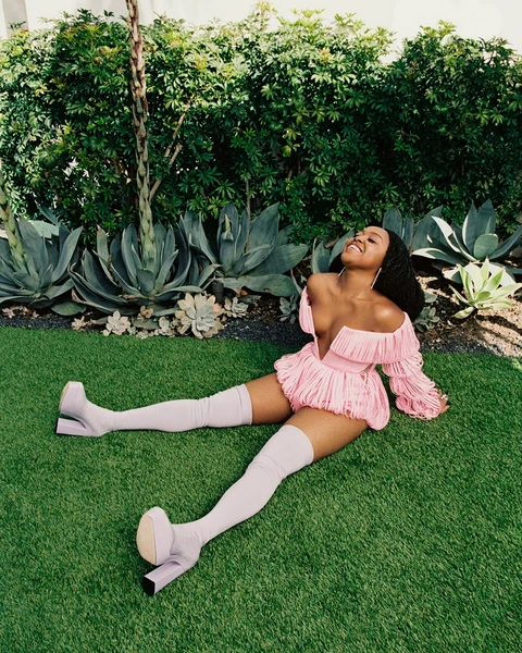 A smiling African American woman in a pink outfit and thigh-high boots lounges on green grass in a garden setting with lush greenery.