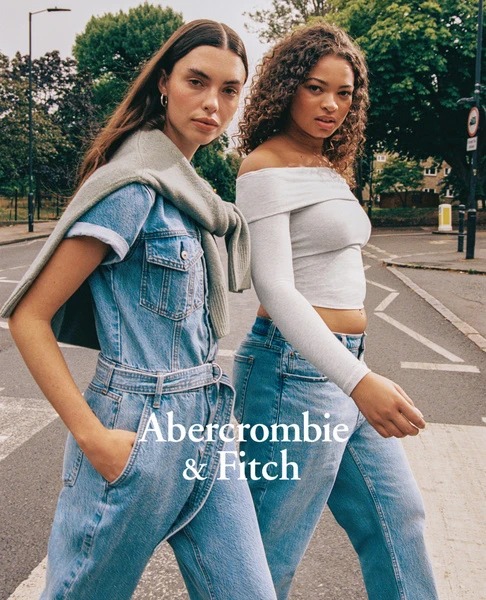Two women in denim outfits walk on a city street, posing for Abercrombie & Fitch. One wears a denim jumpsuit with a light sweater.
