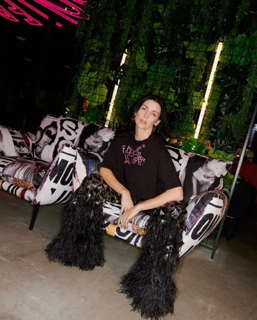 A fashionably dressed adult woman sits on a patterned couch, wearing a black t-shirt and decorative feathered pants in an indoor setting.