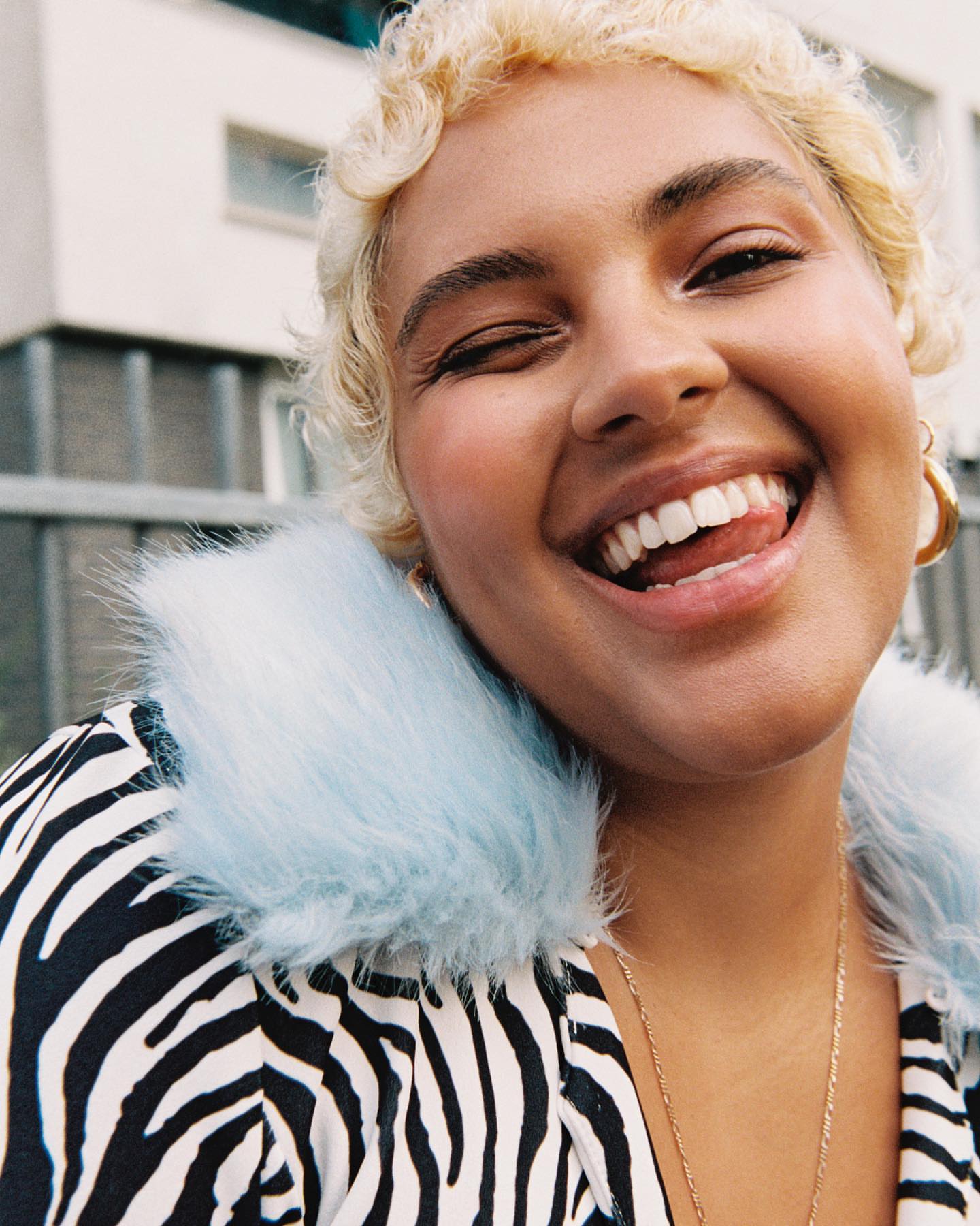 Close-up shot of a woman with short, light-blonde hair, wearing a zebra-print jacket with a blue furry collar, smiling, and winking.