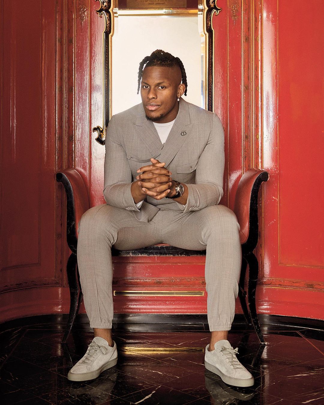 Adult male sits in a decorative chair wearing a grey suit, sneakers and a watch, set against an elaborate red doorway in an indoor setting.