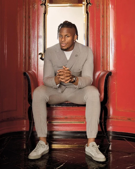 Adult male sits in a decorative chair wearing a grey suit, sneakers and a watch, set against an elaborate red doorway in an indoor setting.