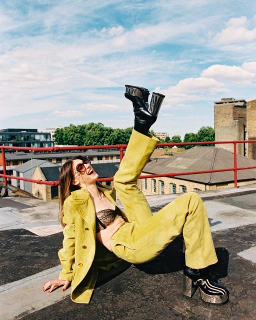 A woman in a yellow suit and black boots poses playfully on a rooftop with city buildings and a bright blue sky in the background.