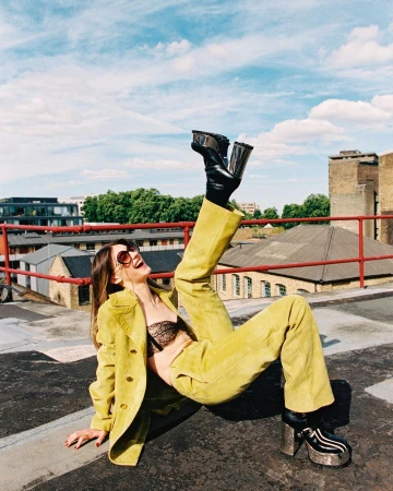A woman in a yellow suit and black boots poses playfully on a rooftop with city buildings and a bright blue sky in the background.