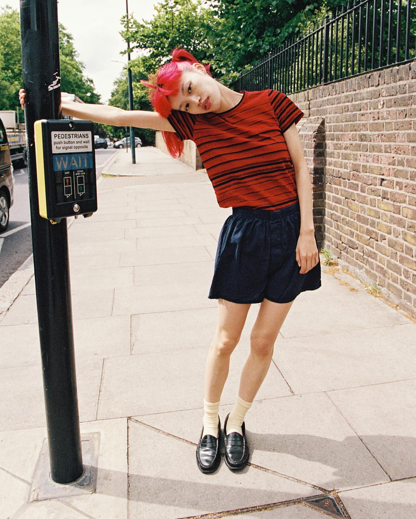 Young adult female with pink hair in casual street style leaning against a pedestrian crossing signal on an urban sidewalk.
