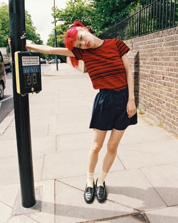 Young adult female with pink hair in casual street style leaning against a pedestrian crossing signal on an urban sidewalk.