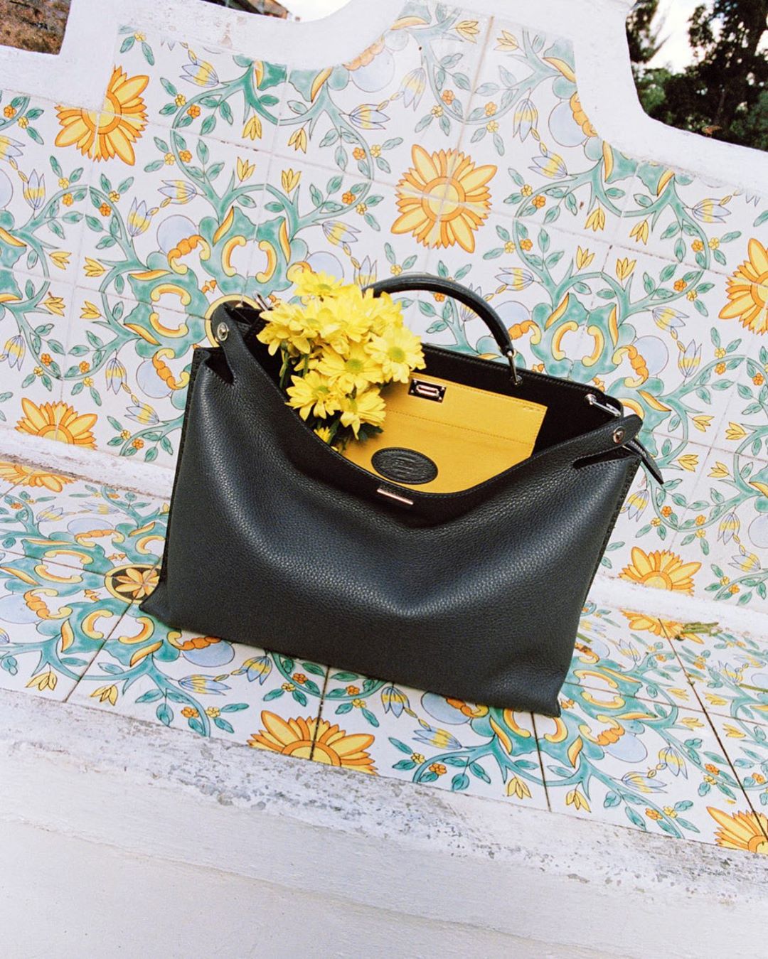 Dark green leather handbag with yellow flowers and a yellow wallet on a patterned bench with light and shadows, fashion still life.