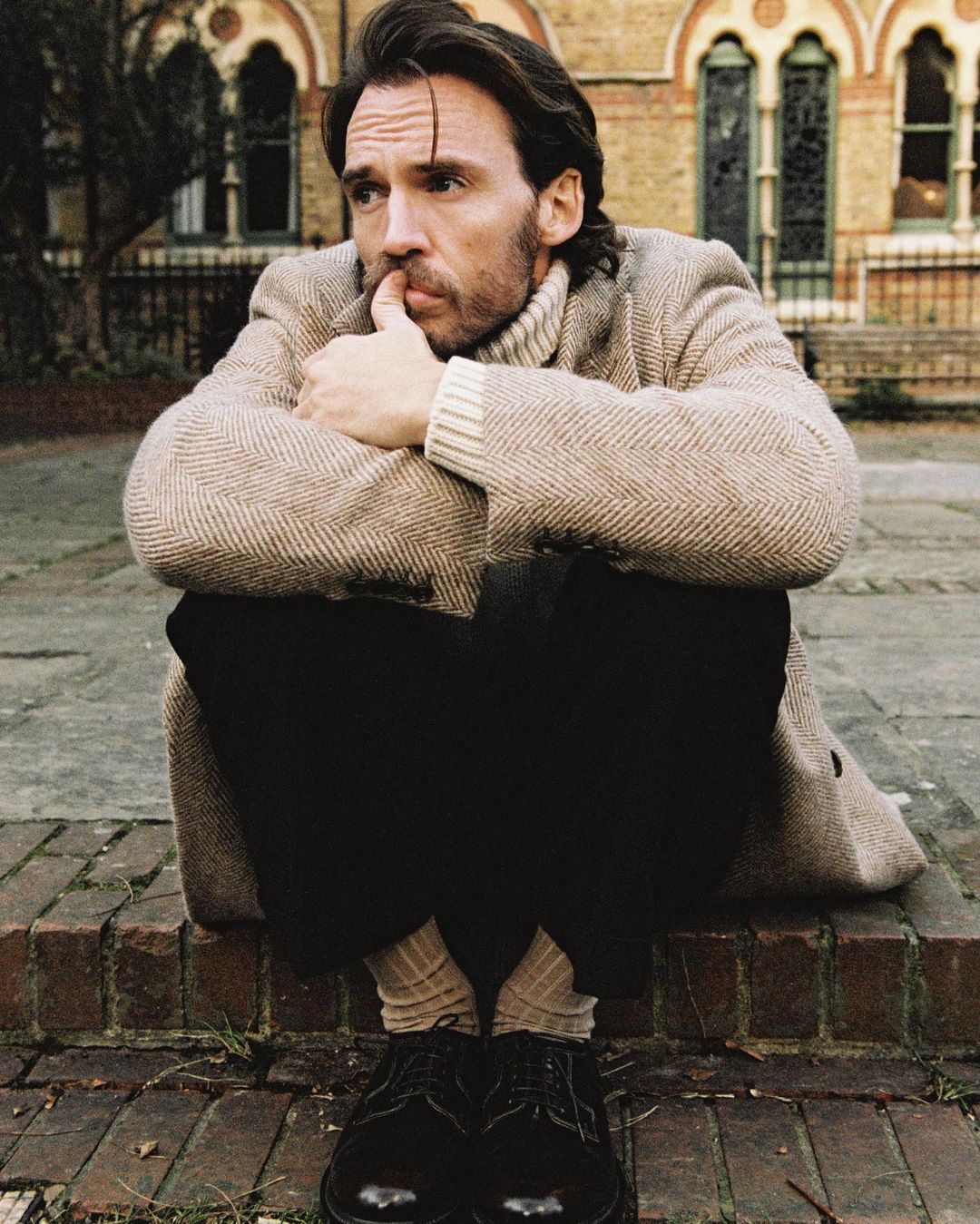 A man in a coat sits on a brick ledge in front of a building, resting his face on his hand in a contemplative pose, looking off to the side.