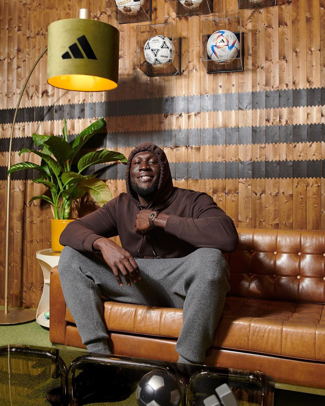 Man in brown hoodie and gray sweats sitting on a brown leather couch indoors with soccer balls and an Adidas lamp.