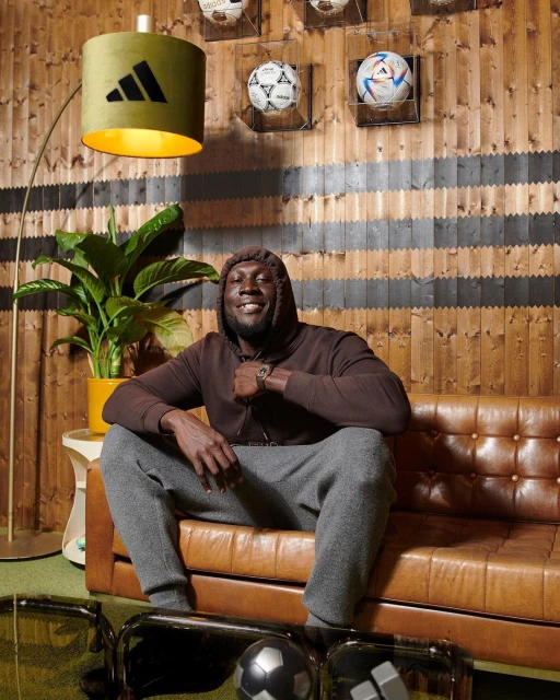 Man in brown hoodie and gray sweats sitting on a brown leather couch indoors with soccer balls and an Adidas lamp.