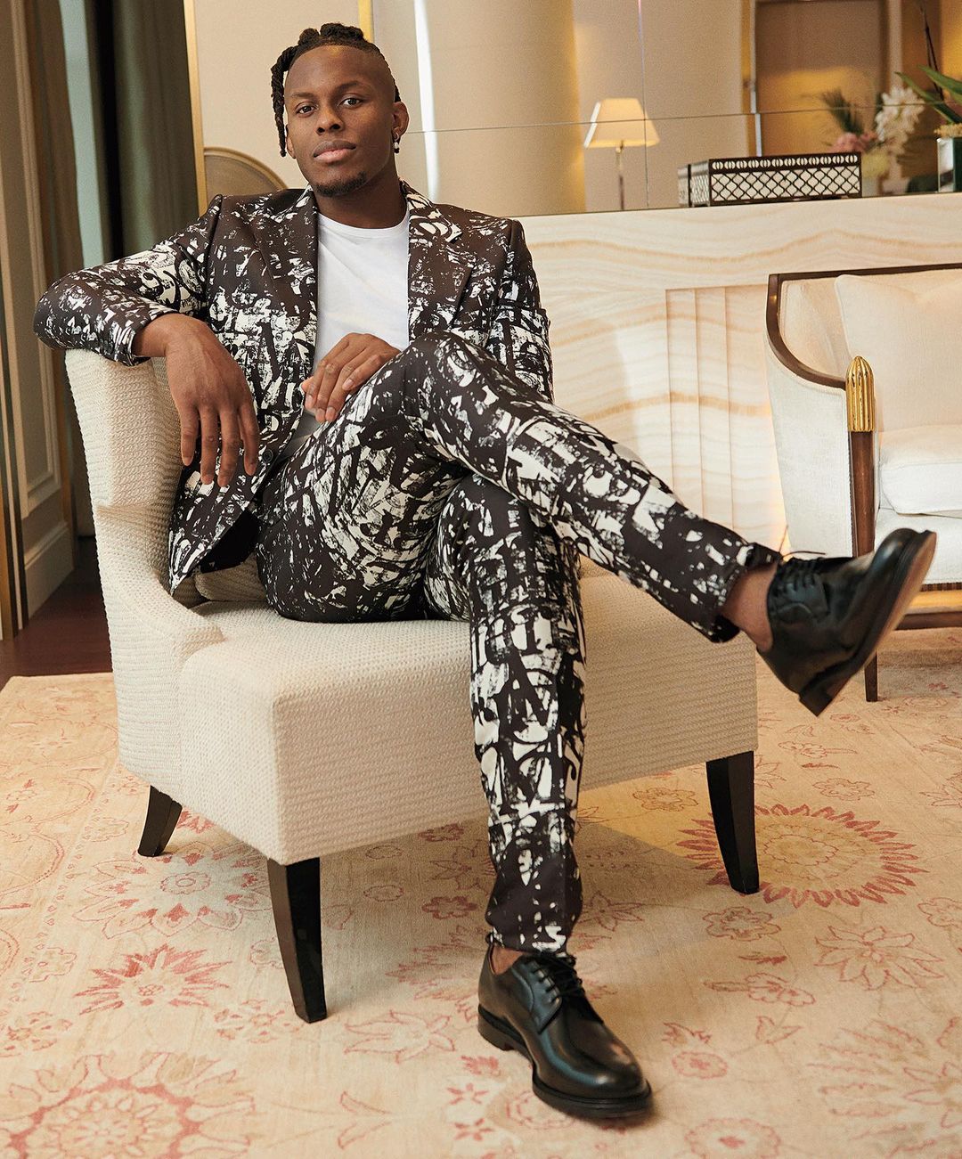 Stylish man in patterned suit sits in a chair indoors. The image captures a blend of fashion and interior design in a relaxed setting.