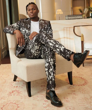 Stylish man in patterned suit sits in a chair indoors. The image captures a blend of fashion and interior design in a relaxed setting.