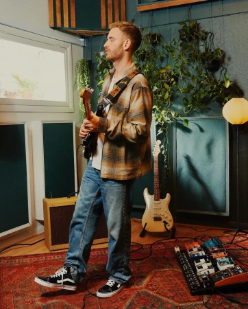 A man holding an electric guitar in a room with indoor plants, music equipment, and a patterned rug, creating a relaxed, musical atmosphere.