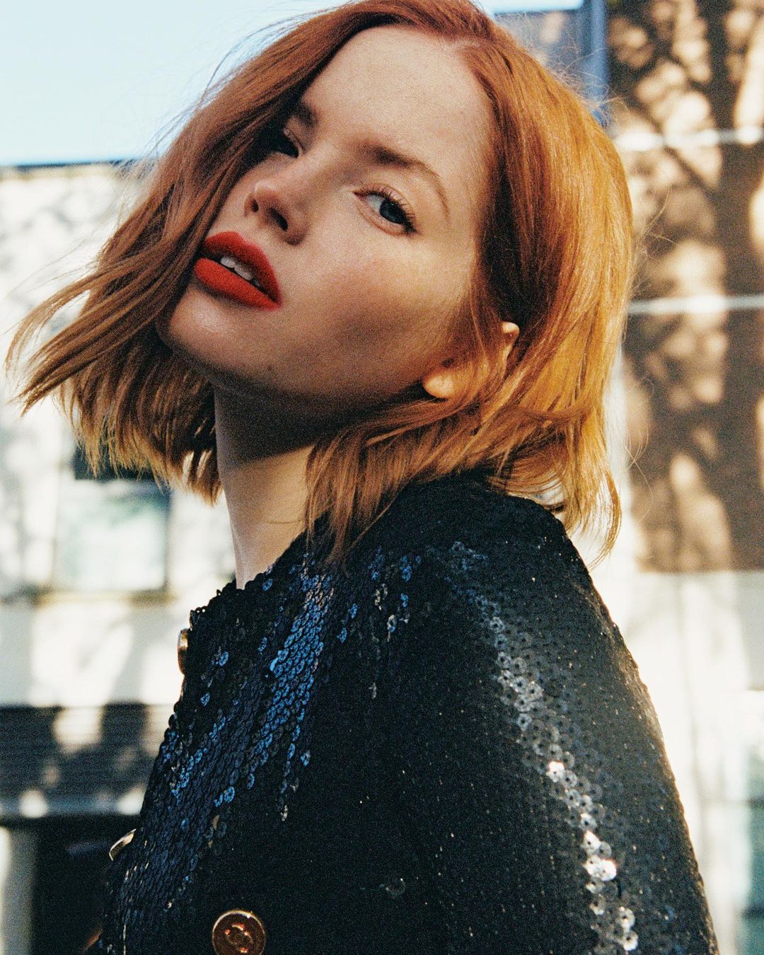 Portrait of a woman with red hair and bright red lipstick wearing a sequined jacket, posing outdoors in natural daylight.