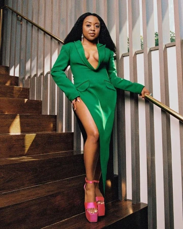 Woman in a green dress and pink platform heels standing on a wooden staircase holding a gold handrail.
