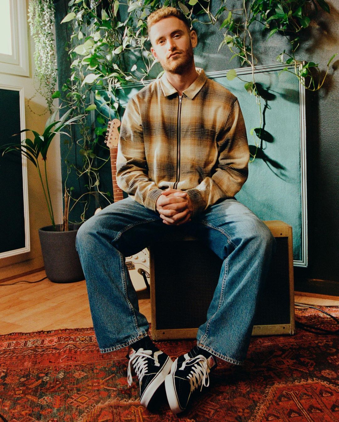A fair-skinned man with red hair and a plaid shirt sits indoors surrounded by plants, a guitar, and music equipment.